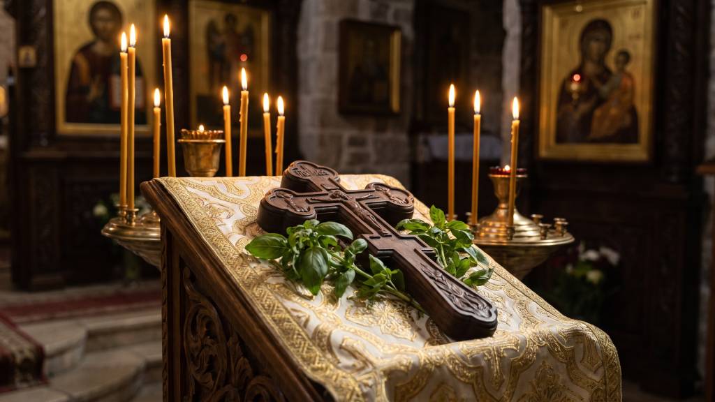 A carved wooden cross decorated with sprigs of fresh basil, resting on an embroidered cloth on a lectern, surrounded by lit beeswax candles in a dim church interior.
