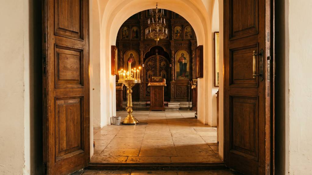 Open wooden doors looking from the narthex into an Orthodox nave, a brass candle stand lit with beeswax candles before the iconostasis.