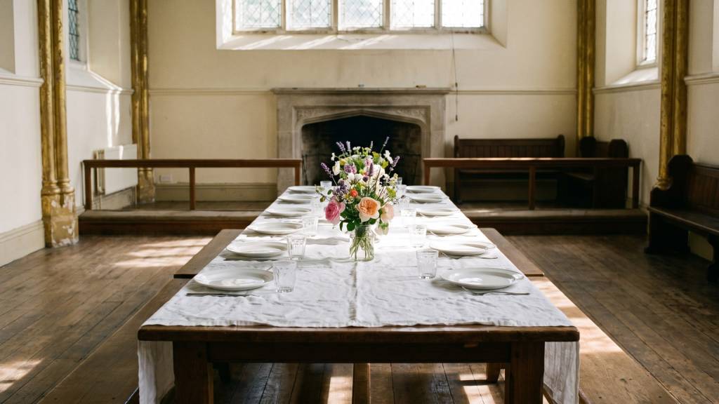 A long parish hall table laid with a white cloth, plates and glasses, and a vase of garden flowers, lit by high windows.