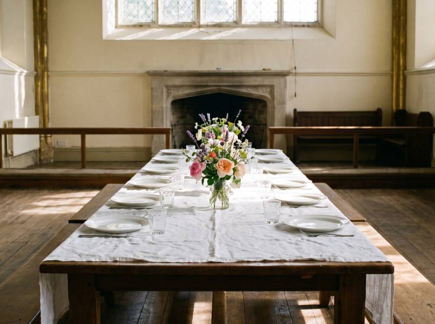 A long parish hall table laid with a white cloth, plates and glasses, and a vase of garden flowers, lit by high windows.