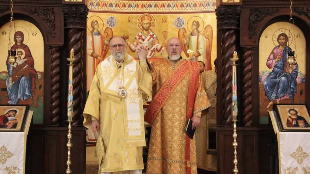 Metropolitan SABA, in gold vestments, raises the hand of newly ordained Deacon Lazarus Nichols, in red and gold vestments, before the iconostasis after the ordination.
