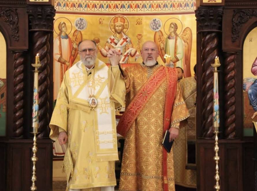Metropolitan SABA, in gold vestments, raises the hand of newly ordained Deacon Lazarus Nichols, in red and gold vestments, before the iconostasis after the ordination.