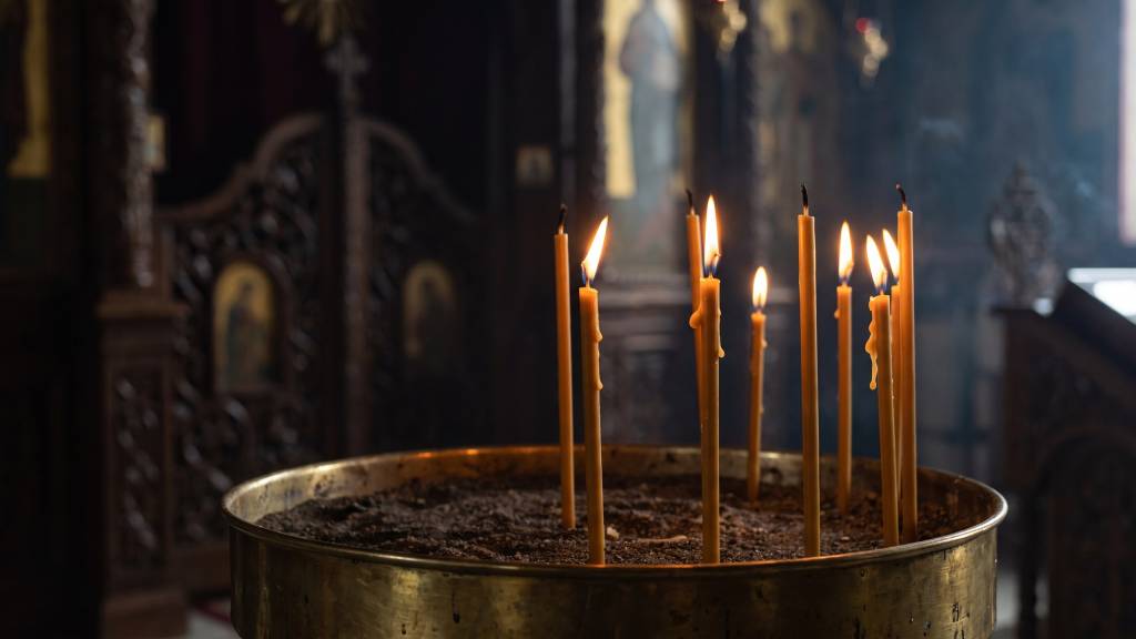Thin beeswax candles burning in a brass sand tray inside a dim Orthodox church.